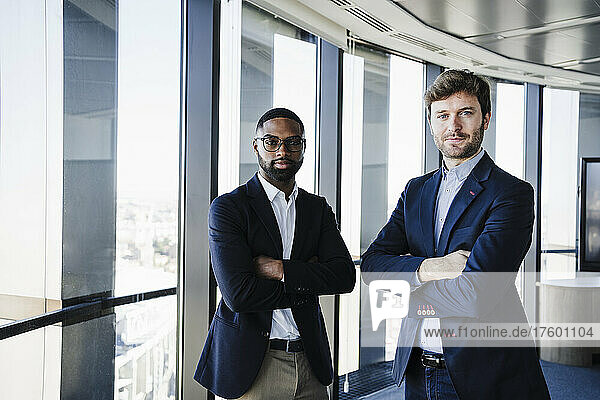 Confident businessmen standing with arms crossed by glass window in office
