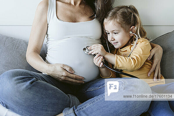 Girl listening to pregnant mother's belly through stethoscope at home