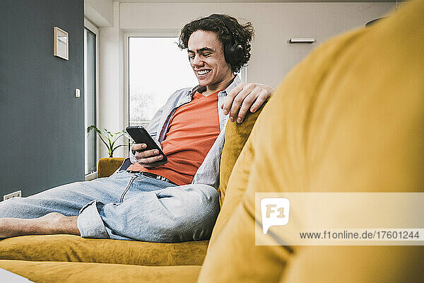 Cheerful man listening music through wireless headphones using smart phone in living room