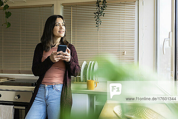 Thoughtful woman with smart phone in kitchen at home