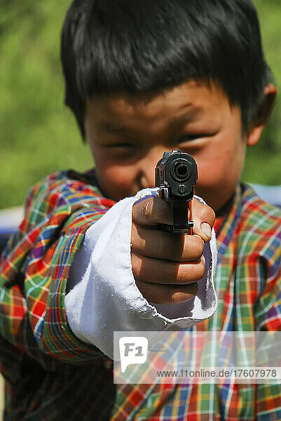 A young boy dressed in traditional clothing points a toy gun at the camera; Paro  Bhutan