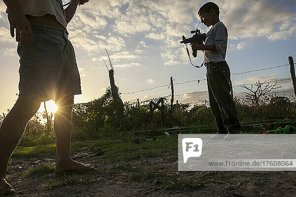 Während die Sonne hinter einem Stacheldrahtzaun untergeht  hält ein kleiner Junge ein Spielzeuggewehr; Cienfuegos  Kuba