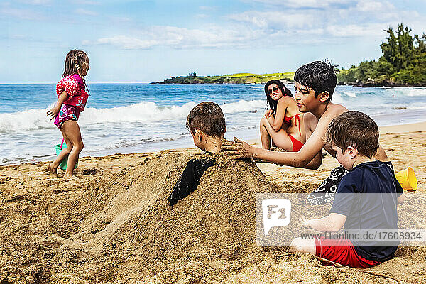 Mother sitting on D. T. Fleming Beach at the water's edge and watching her children playing and burying a sibling in the sand; Kapalua  Maui  Hawaii  United States of America