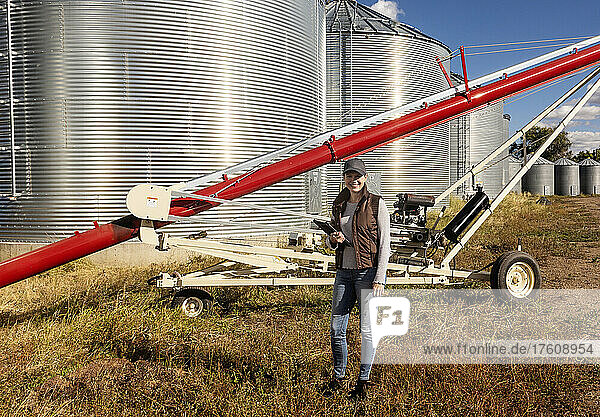 Porträt einer reifen Frau  die auf ihrer Farm arbeitet  einen Tablet-Computer in der Hand hält und vor Getreidebehältern und einer Schnecke steht; Alcomdale  Alberta  Kanada