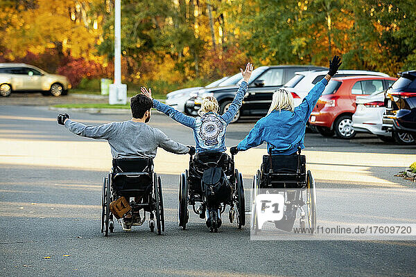 Drei junge Querschnittsgelähmte in ihren Rollstühlen  die an einem schönen Herbsttag mit erhobenen Armen in einer Reihe über einen Parkplatz in einem Park fahren; Edmonton  Alberta  Kanada
