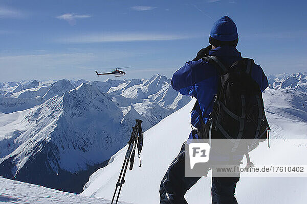 A back country skiier watches a heli-ski chopper approach a peak.; Selkirk Mountains  British Columbia  Canada.