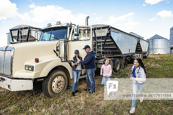 Mature couple working on their farm  standing next to a diesel transport truck and consulting their tablet computer while their two daughters have fun running around next to them; Alcomdale  Alberta  Canada