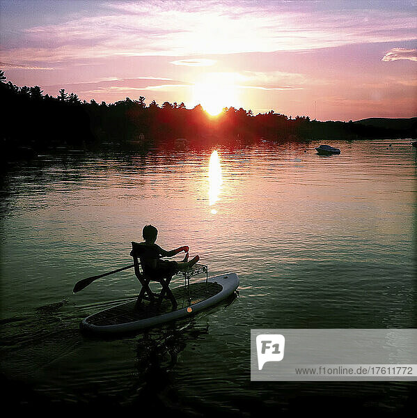Ein Junge versucht  bei Sonnenuntergang auf seinem Standup-Paddle-Board zu sitzen; Sebago Lake  Maine  Vereinigte Staaten.