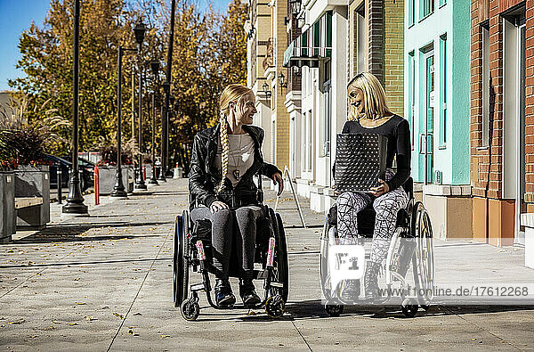 Zwei junge  querschnittsgelähmte Frauen in ihren Rollstühlen  die an einem schönen Herbsttag gemeinsam auf einem Gehweg spazieren gehen  eine hält eine Einkaufstasche; Edmonton  Alberta  Kanada