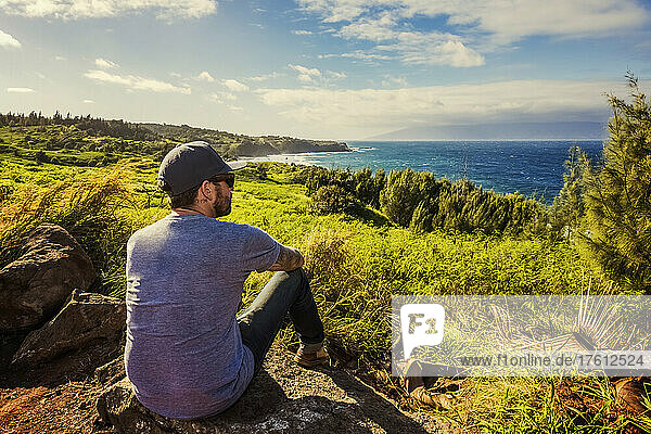 Ein Mann sitzt auf einem Felsen und blickt vom Honoapiilani Hwy aus auf den Ozean und die Küste; Maui  Hawaii  Vereinigte Staaten von Amerika