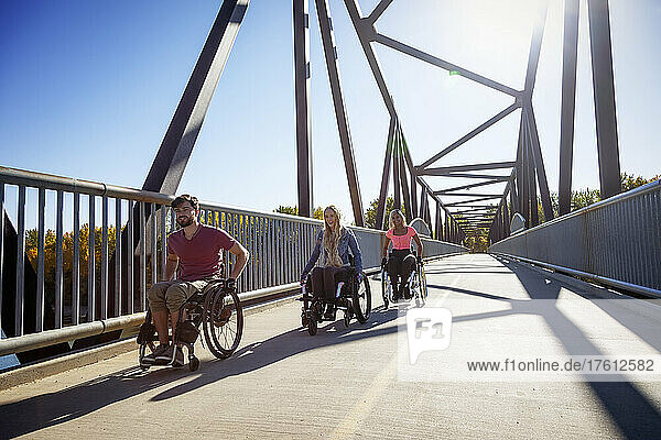 Three young paraplegic friends spending time together moving across a park bridge in their wheelchairs in a city park on a beautiful day; Edmonton  Alberta  Canada