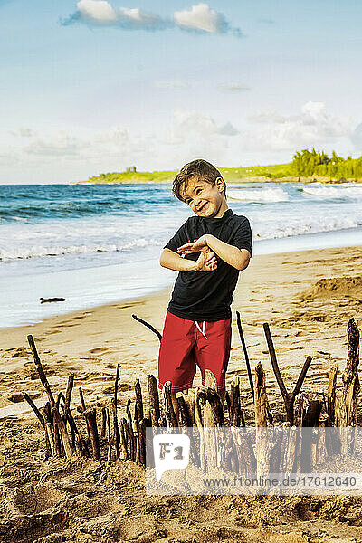 A young boy stands on the beach and proudly displays his creation in the sand at D. T. Fleming Beach; Kapalua  Maui  Hawaii  United States of America