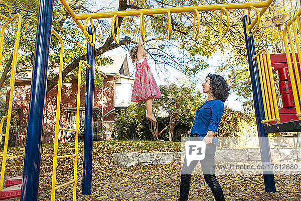 Mother with young daughter at a playground as she watches her daughter hanging from high climbing bars with pride; Toronto  Ontario  Canada
