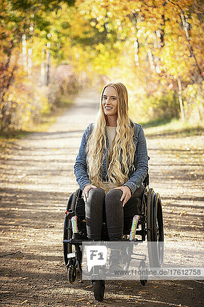 Junge querschnittsgelähmte Frau in ihrem Rollstuhl  die an einem schönen Herbsttag einen Weg in einem Park hinunterfährt; Edmonton  Alberta  Kanada
