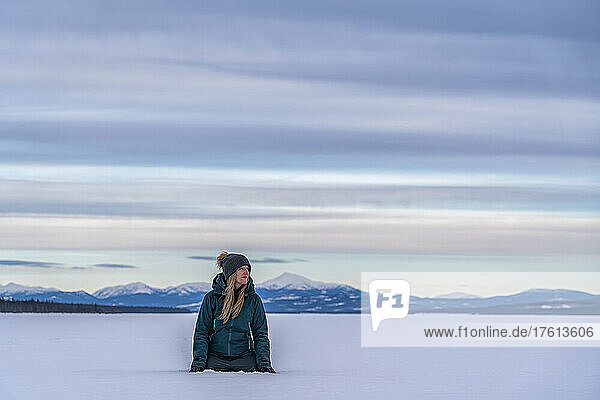 Frau kniet im tiefen Schnee und betrachtet die Landschaft am zugefrorenen Tagish Lake; Tagi; Tagish  Yukon  Kanada