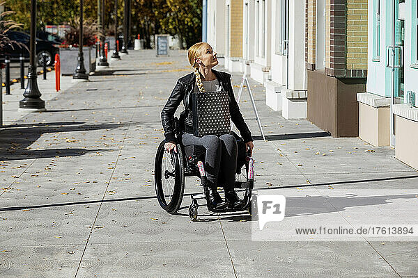 Junge querschnittsgelähmte Frau in ihrem Rollstuhl mit einer Einkaufstasche  die an einem schönen Herbsttag einen städtischen Gehweg hinunterfährt; Edmonton  Alberta  Kanada