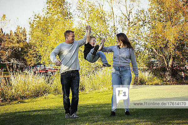 Mutter und Vater schwingen ihren kleinen Sohn zwischen sich  während sie gemeinsam in einem Stadtpark spazieren gehen; St. Albert  Alberta  Kanada
