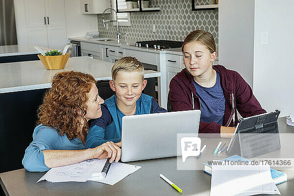 A mother with her teenage daughter and young son sit at the kitchen table at home with a laptop and tablet doing school work while being homeschooled; Alberta  Canada