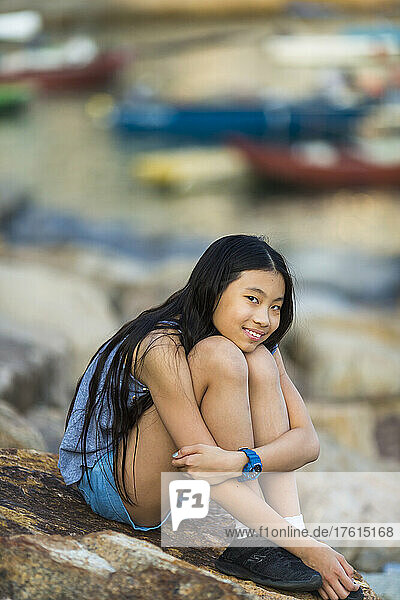 Young girl sits on a rock on the shore of a harbour; Stanley  Hong Kong  China