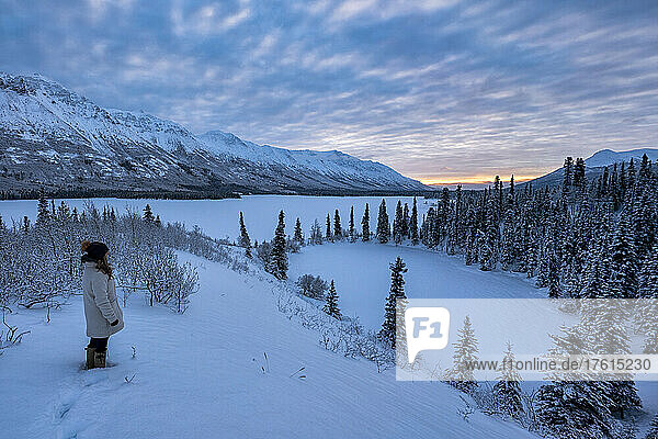 Woman standing in the snow on a mountain top looking out at a stunning landscape with the sun rising over Annie Lake in Winter; Whitehorse  Yukon  Canada