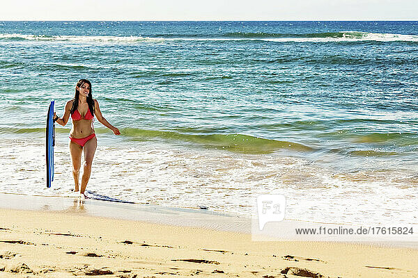 Eine Frau in einem Bikini trägt ein Bodyboard  während sie aus dem Wasser auf den Sand am D. T. Fleming Beach geht; Kapalua  Maui  Hawaii  Vereinigte Staaten von Amerika