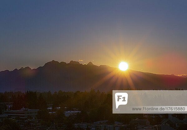 Brillanter Sonnenuntergang hinter den Bergen mit Sonnenstrahlen  die über den Wald und die Stadt darunter fluten; Surrey  British Columbia  Kanada