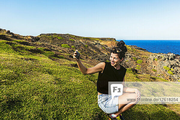 Junge Frau kauert am Ufer von Nakalele Point und macht ein Selbstporträt mit der zerklüfteten Küste der Insel und dem Meer im Hintergrund; Maui  Hawaii  Vereinigte Staaten von Amerika