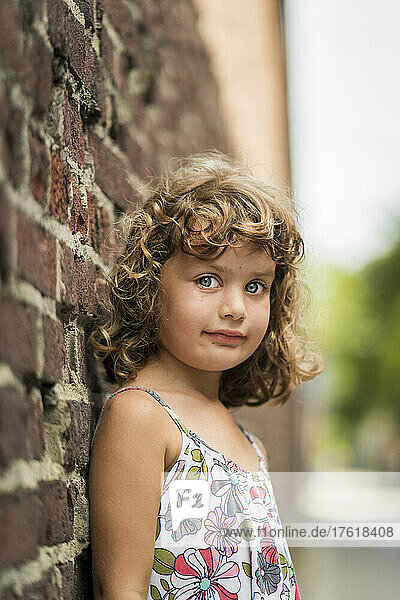 Portrait of a young girl with curly blond hair and blue eyes standing against a brick wall; Toronto  Ontario  Canada