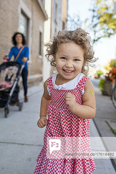Preschooler girl running down a sidewalk in her red and white gingham dress and looking into the camera with her mother and a stroller in the background; Toronto  Ontario  Canada