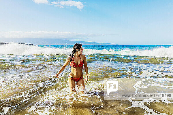 Frau im roten Bikini steht im Meerwasser vor der Küste und spielt in den Wellen am D. T. Fleming Beach mit der Insel Molokai in der Ferne; Kapalua  Maui  Hawaii  Vereinigte Staaten von Amerika