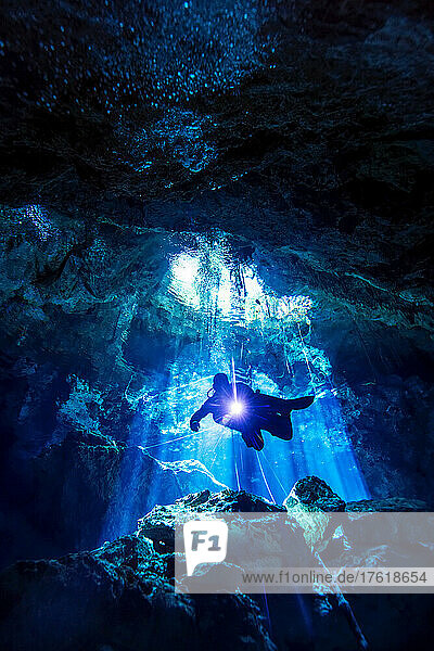 Ein Höhlentaucher steigt in ein Höhlensystem hinab  während im Hintergrund Lichtstrahlen das Wasser durchdringen.