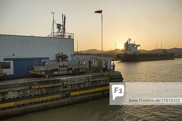 An den Pedro-Miguel-Schleusen stehen und arbeiten mehrere Männer entlang des Panamakanals und der Ausrüstung  während die Sonne hinter einem großen Schiff untergeht; Panama