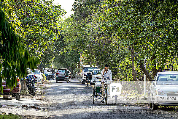 Street scene in a village in India with vehicles and motorcycles  and a man cycling with a trailer and talking on the phone; Greater Noida  Uttar Pradesh  India