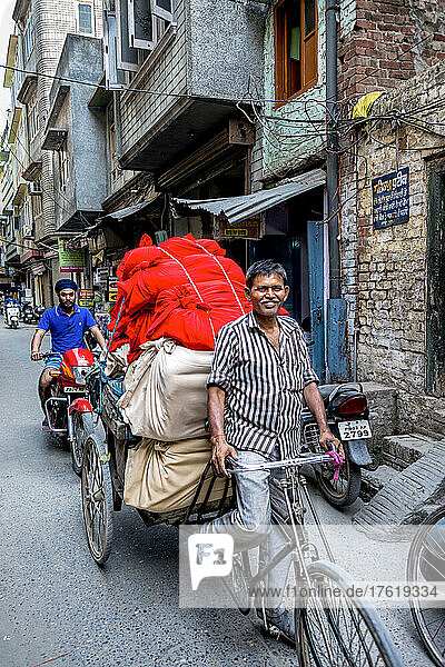 Man rides a bicycle pulling a loaded trailer behind  with a man on a motorcycle following behind on a street in India; Amritsar  Punjab  India