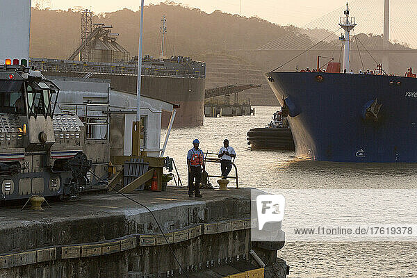 An den Pedro-Miguel-Schleusen  in der Nähe der Centennial Bridge  stehen und arbeiten mehrere Männer neben dem Panamakanal  der Ausrüstung und den großen Schiffen in Panama; Panama