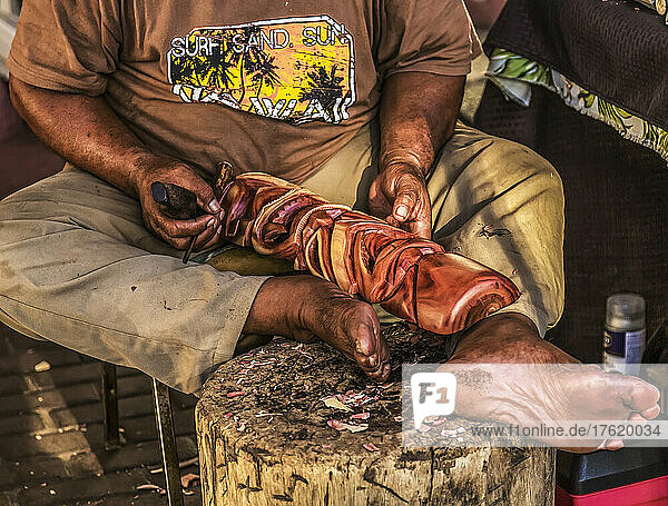 Ein hawaiianischer Mann sitzt barfuß und schnitzt mit einem Werkzeug Holz  um ein traditionelles Kunsthandwerk herzustellen; Maui  Hawaii  Vereinigte Staaten von Amerika