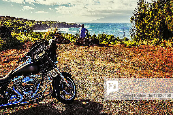 A man sits on a rock looking out at the ocean with the Island of Molokai in the distance and with his motorcycle parked behind him at a scenic viewpoint on the Honoapiilani Highway; Maui  Hawaii  United States of America