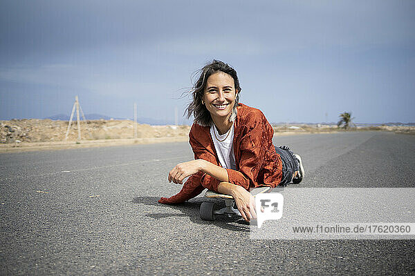 Smiling young woman lying over skateboard on road