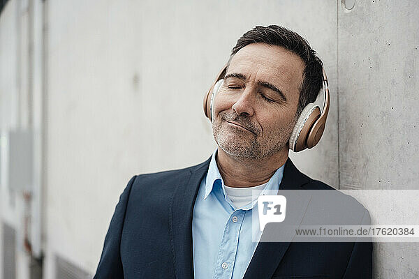 Lächelnder Geschäftsmann mit geschlossenen Augen  der Musik über drahtlose Kopfhörer an der Wand hört