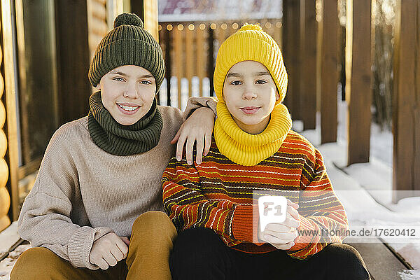 Boys wearing warm clothing and knit hat sitting at porch of house in winter