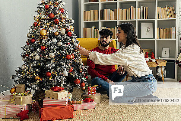 Young man sitting by girlfriend decorating Christmas tree at home