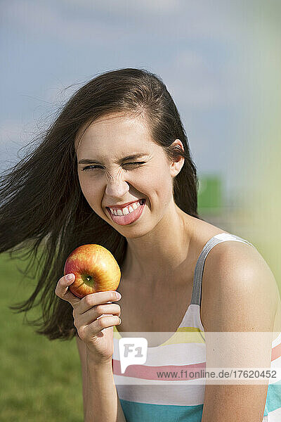 Young woman with apple winking eye and sticking out tongue on sunny day