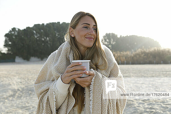 Smiling woman in blanket holding tea cup at beach