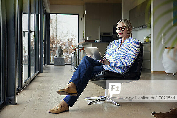 Frau mit Brille und Tablet-PC sitzt zu Hause auf einem Stuhl
