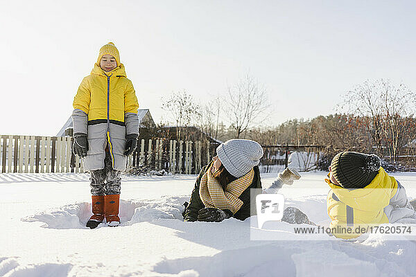 Woman with son lying on snow looking at boy standing in winter
