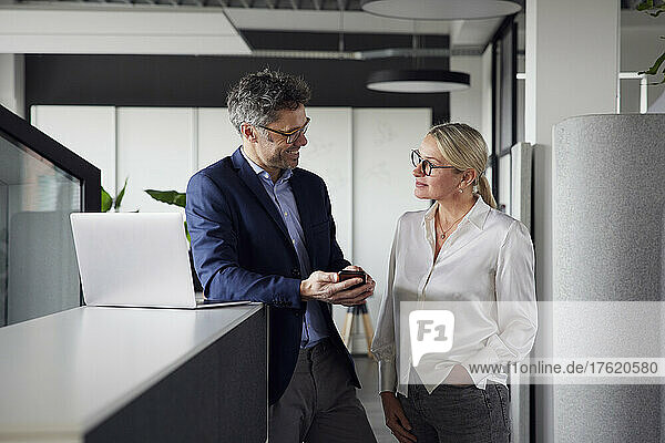 Geschäftsfrau blickt mit der Hand in der Tasche auf einen Kollegen  der im Büro sein Smartphone hält