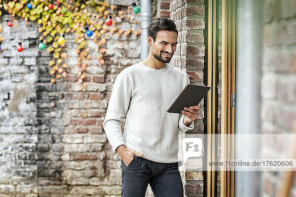 Happy freelancer with hand in pocket using tablet PC by brick wall