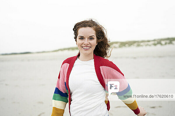 Glückliche rothaarige Frau in buntem Cardigan-Pullover  die Spaß am Strand hat