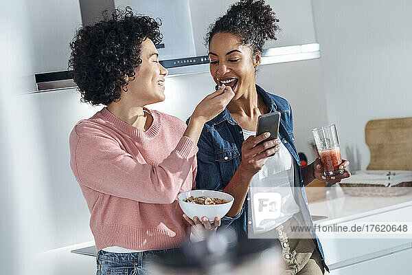 Young woman feeding cereal to friend standing in kitchen at home