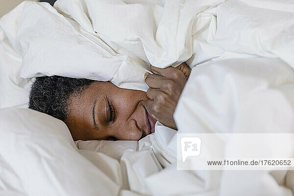 Lächelnde Frau liegt zu Hause auf dem Bett  das mit einer weißen Bettdecke bedeckt ist